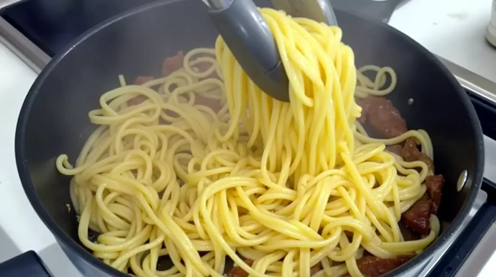 Tongs tossing noodles and steak in a skillet with garlic butter